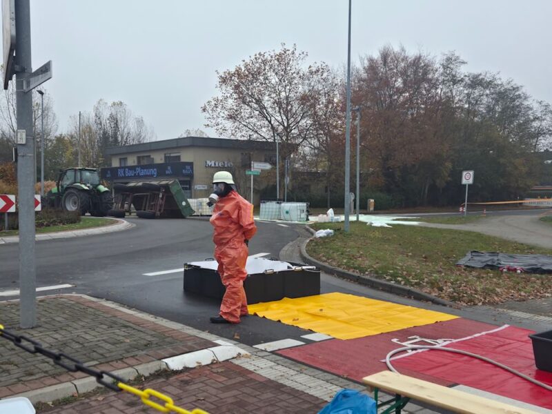 In Schutzausrüstung: Einsatzkraft an der vorbereiteten Dekonstelle. Im Hintergrund der verunfallte Anhänger sowie die ausgelaufene Beladung. (Foto: Kreisfeuerwehr Rotenburg (Wümme))